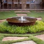 Round metal water feature with fountain surrounded by grass and flagstone path