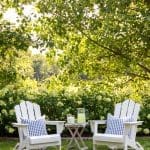 Two white Adirondack chairs with blue gingham pillows on a lawn, under trees.