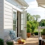White siding exterior with open French doors, white bench with blue pillow, basket of flowers, and