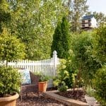 Wicker chair with blue floral pillow near white picket fence and birdhouse