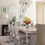 White dining room with a gold chandelier, floral artwork, and a black table.