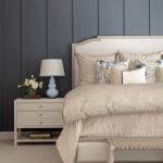 Gray paneled headboard with beige bedding and a lamp.