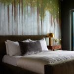 Dark green wall with a forest mural, bed with gray pillows and a gold lamp.