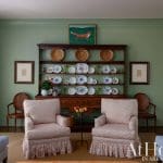 Green wall, wooden shelving with plates, and two patterned chairs.