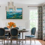 White dining room with blue chairs, a floral rug, and a landscape painting.