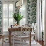 White dining table with wicker chairs, window shutters, and patterned wallpaper.