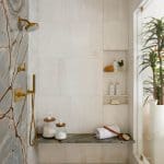 Shower with marble-look tile, gold fixtures, stone bench, and built-in shelves.