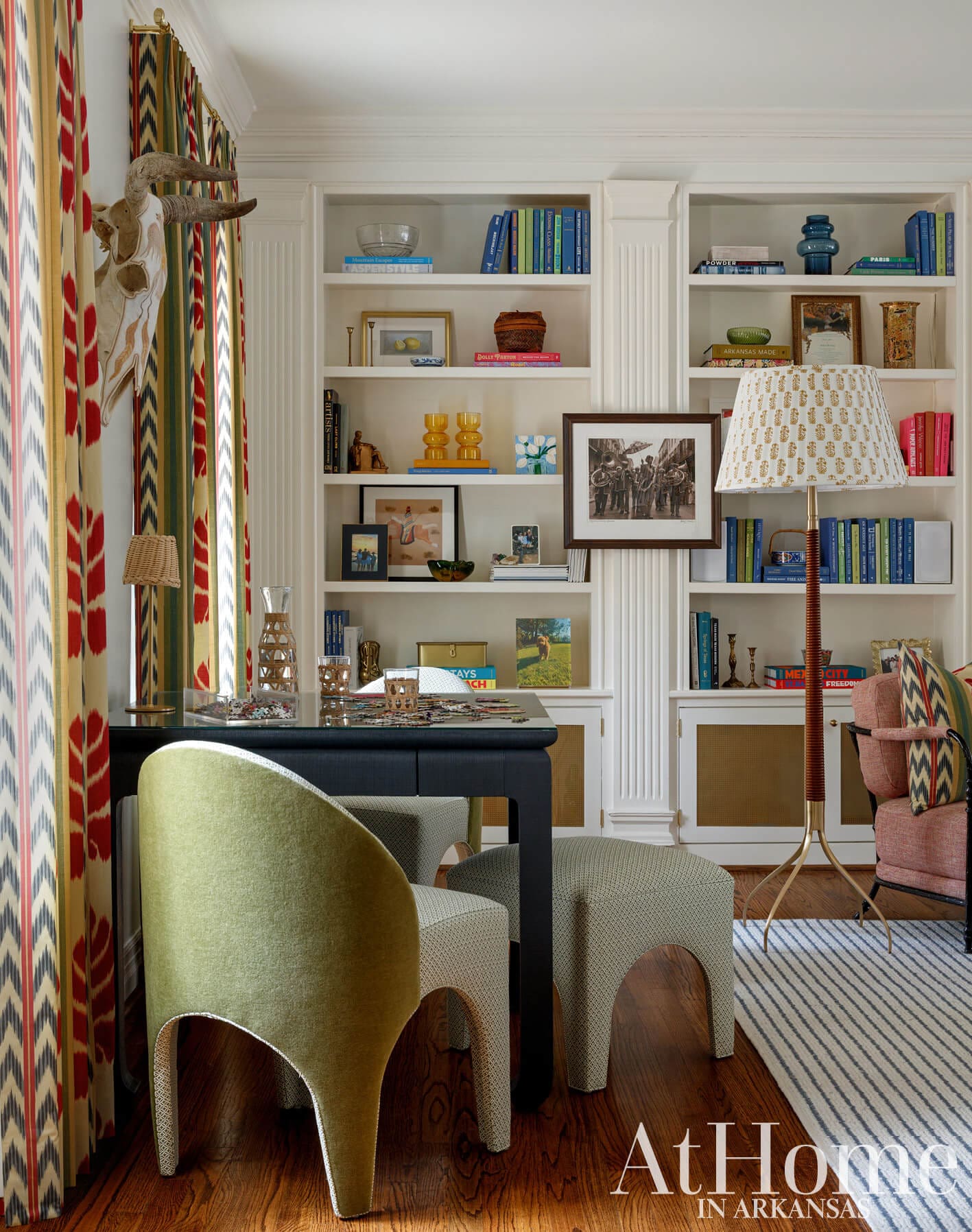 Green upholstered chair at desk with puzzle, flanked by patterned curtains and built-in bookshelves.