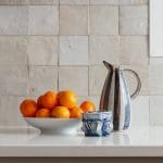 Kitchen counter with bowl of oranges, ceramic pitcher, and blue vase against tiled backsplash.