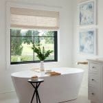 White freestanding bathtub with brass faucet, natural woven shade, and abstract art.