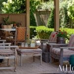 Covered porch with wooden chairs and tables, striped cushions, and lush greenery.