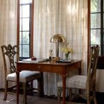 Wooden desk with two chairs, brass lamp, and bust in front of grid-patterned curtains.
