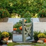 Green garden gate with pumpkins and mums, stone pathway, and brick wall
