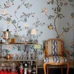Bar cart with glassware and bottles in front of floral wallpaper and upholstered chair.