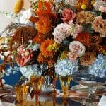 Floral arrangement with orange, pink, and blue flowers on a table with amber glasses