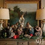 Nativity scene with figurines and greenery on a wooden surface, flanked by lamps.