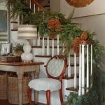 Staircase decorated with pine garland and pinecones, featuring a striped lamp and antique chair.