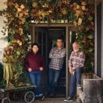 Three men standing on a porch decorated with a large garland and bows.