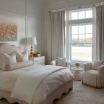 Bedroom with pink scalloped bed, floral art, neutral bedding, and two armchairs by window.