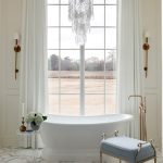 White freestanding bathtub with brass faucet, blue upholstered bench, and large window view.