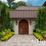 Luxury home entrance with arched wooden double doors, stone pathway, and manicured landscaping.