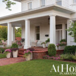 White house with wrap-around porch, brick walkway, and potted flowers.