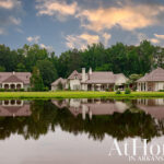 Three white houses with brown roofs reflected in a pond, surrounded by green trees.