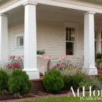 White porch with wicker furniture, pink cushions, and flowering plants.