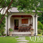 Covered outdoor kitchen with stone wall, wooden cabinets, and dining set. Lush garden and trees