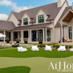 Modern farmhouse with stone chimney, white siding, and dark windows. Backyard features putting