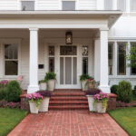 White house with red brick path, porch with columns, and potted flowers.