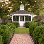 White gazebo with black trim and cupola, surrounded by manicured hedges and brick pathway.