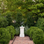Brick pathway leading to a sundial on a pedestal, surrounded by manicured bushes and trees.