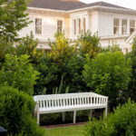 White slatted bench in garden with white house and lush green foliage