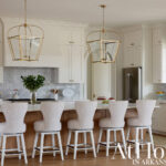 White kitchen with marble backsplash, brass pendant lights, and upholstered bar stools.