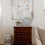 Abstract art above a blue and white lamp on a marble-top dresser. White stool nearby.