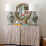 Two ginger jar lamps flank a round mirror above a striped skirted console table.