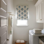 Laundry room with white walls, blue floral roman shade, white washer and dryer, and wicker basket