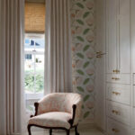 Pink floral armchair next to floral wallpaper and white cabinets.