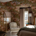 Bedroom with bird and tree patterned wallpaper, upholstered chair, and gold luggage rack.