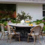 Oval dining table with wicker base and chairs on gravel patio, white brick house background