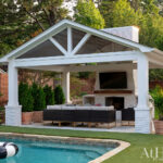 Covered outdoor living space with fireplace, TV, and seating by a pool.