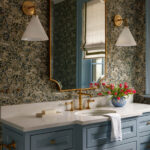 Blue vanity with marble countertop, brass fixtures, and floral wallpaper