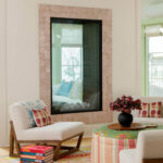 Living room with pink tile fireplace, boucle chairs, patterned ottoman, and stack of colorful books.