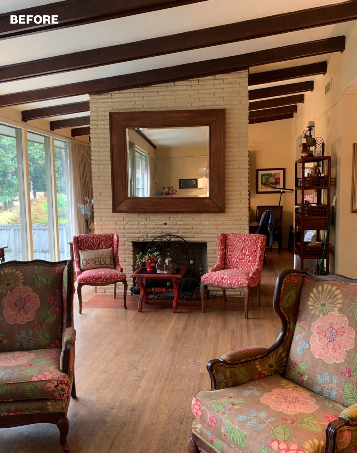 Living room with floral upholstered chairs, a brick fireplace, and wood-beamed ceiling.