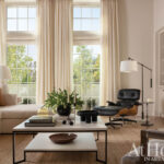 Living room with sectional sofa, Eames lounge chair, and large windows with cream curtains.