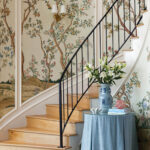 Staircase with floral wallpaper, wood treads, and black railing. Blue table with vase.