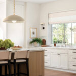 Kitchen island with marble countertop, light wood base, and bar stools. White cabinets and window