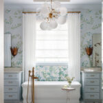 Light blue bathroom with floral wallpaper, freestanding tub, brass fixtures, and glass chandelier.