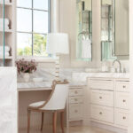 Cream bathroom vanity with marble countertop, ornate chair, and shelves with towels.